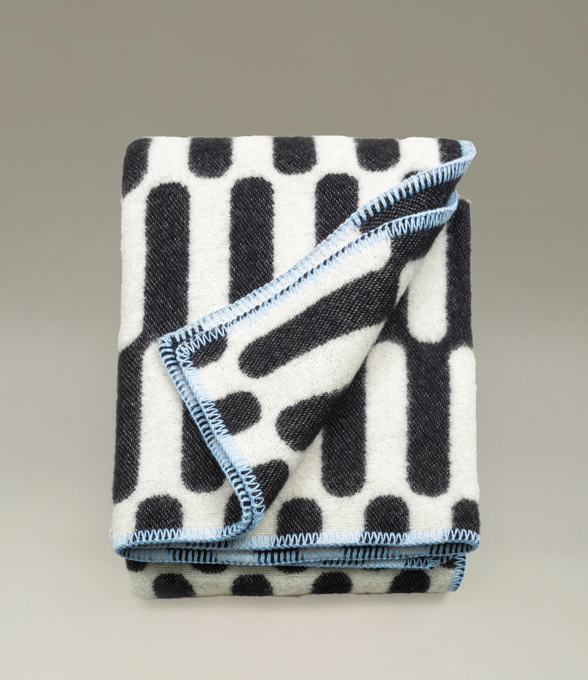 Folded monochrome wool blanket featuring a bold black and white geometric pattern with vertical bars and dot accents. A contrasting light blue stitched border adds a subtle pop of color, neatly finishing the design. The blanket is displayed against a neutral background to emphasize its modern aesthetic.