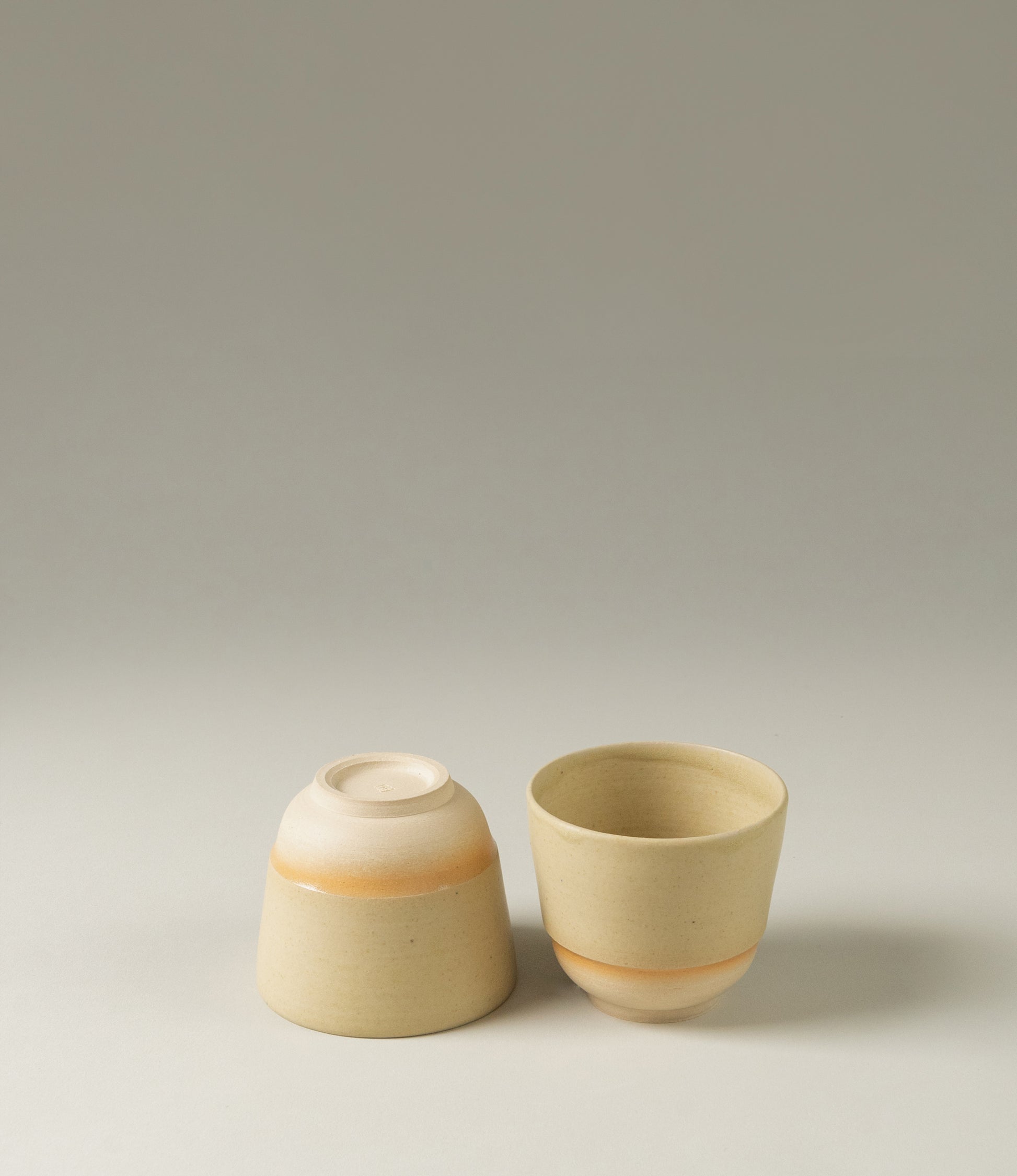 A cup set from Omni Ceramics. The handmade pieces have a soft orange painted gradient ring on them. The item is coming in a light natural beige color.